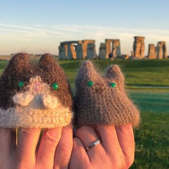 A tuxedo kitty mini and a grey kitty mini, both made with real cat fur, held up in front of Stonehenge | Kitten KaZoedle